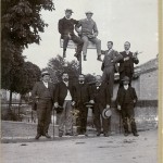 Vorne stehen fünf Personen, zwei stehen auf der Mauer, und zwei weitere sitzen auf einem Wegweiser. Der Wegweiser verweist nach links: ..wies 3,7 und Aspelt 7 km und nach rechts Diedenhofen 21 km. Alle Männer bis auf einen haben einen Hut, sei es auf dem Kopf oder in der Hand, und einen Spazierstock.