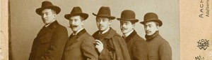 5 Männer im Mantel, hintereinander stehend mit Hut.