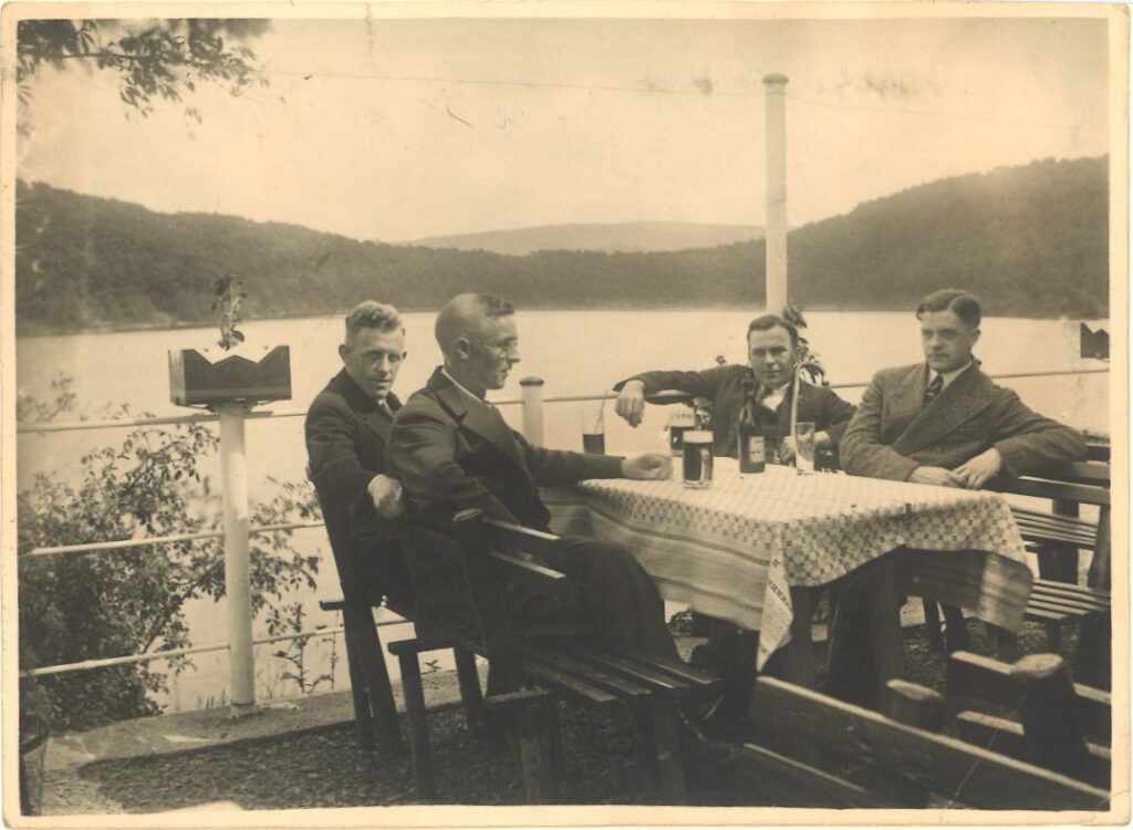 Vier Männer auf der Terrasse eines Lokals an einem Tisch mit Biergläsern. Im Hintergrund ein See. Vielleicht der Rursee.