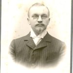Mann mit Spitzbart und Nickelbrille im gestreiftem Anzug mit Stehkragen
